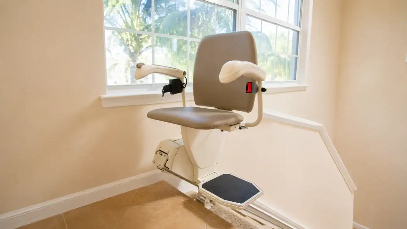 A stair lift chair with armrests and a footrest is installed on a staircase rail inside a beige, sunlit room with a window.