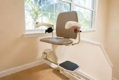 A stair lift chair with armrests and a footrest is installed on a staircase rail inside a beige, sunlit room with a window.