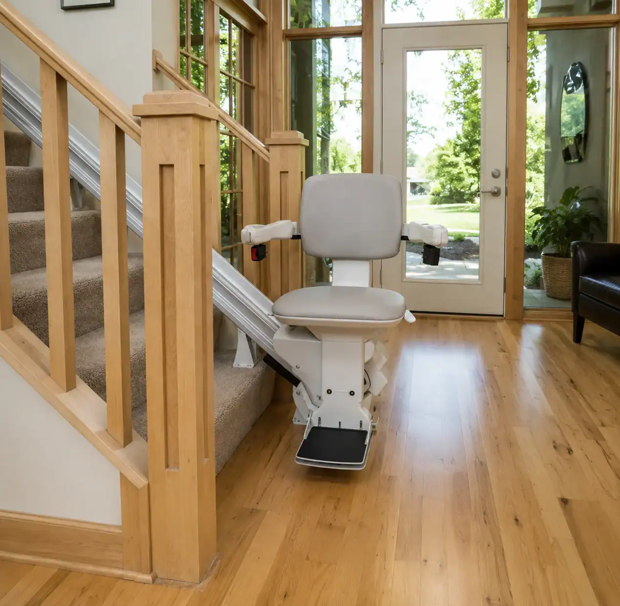 A stairlift is installed on a carpeted staircase next to a wooden handrail in a home with wooden floors and large windows.