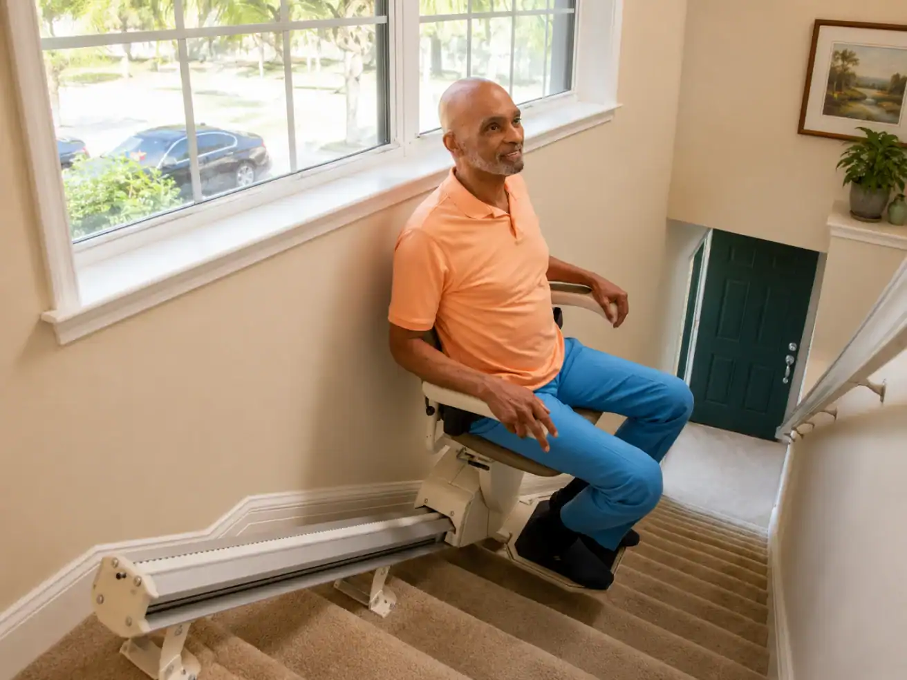 A man sits on a stairlift going up a carpeted staircase in a well-lit home with large windows and potted plants visible.