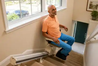 A man sits on a stairlift going up a carpeted staircase in a well-lit home with large windows and potted plants visible.