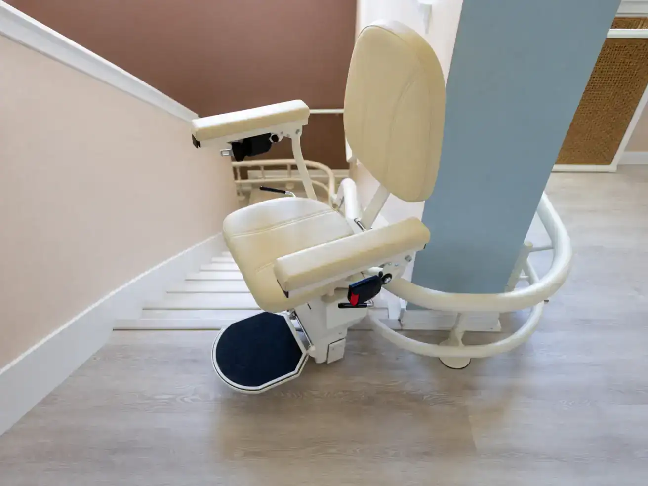 A beige stairlift chair is installed on a staircase with light wood flooring and cream-colored walls, designed to assist mobility-impaired individuals.