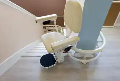 A beige stairlift chair is installed on a staircase with light wood flooring and cream-colored walls, designed to assist mobility-impaired individuals.