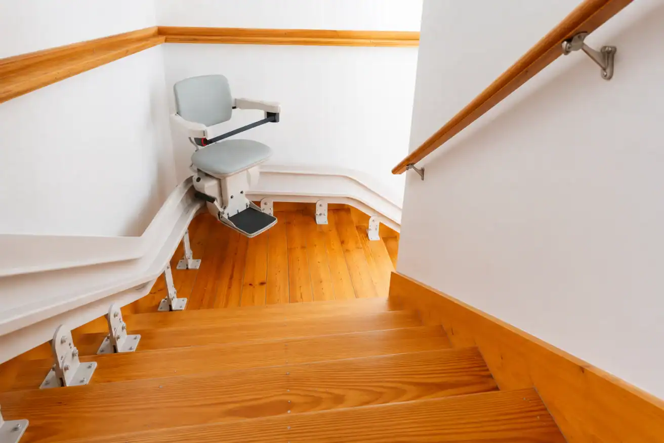 A stairlift chair is installed on a wooden staircase with a curved rail system, shown at the landing and surrounded by white walls with wooden trim.