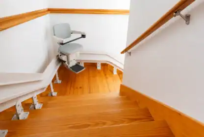 A stairlift chair is installed on a wooden staircase with a curved rail system, shown at the landing and surrounded by white walls with wooden trim.