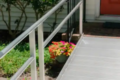 A metal wheelchair ramp with railings leads to a house entrance with a red door, surrounded by plants and flowers.