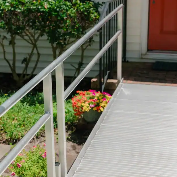 A metal wheelchair ramp with railings leads to a house entrance with a red door, surrounded by plants and flowers.