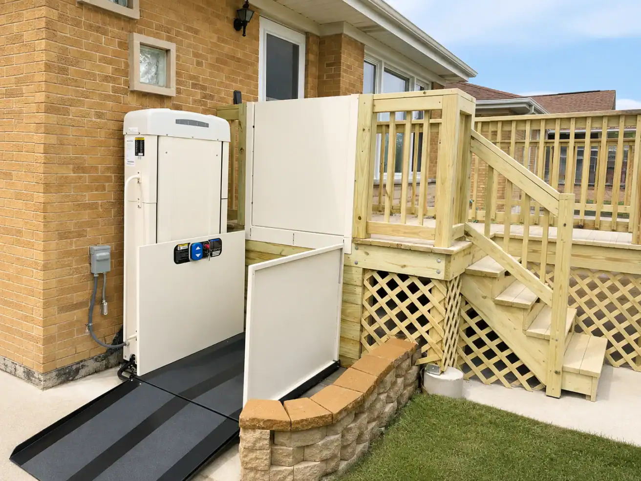A vertical platform lift is installed next to a wooden porch, providing wheelchair access from the yard to the elevated entrance of a brick house.