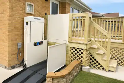 A vertical platform lift is installed next to a wooden porch, providing wheelchair access from the yard to the elevated entrance of a brick house.