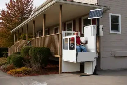 A man operates an outdoor vertical platform lift attached to a porch, with a control panel and a solar panel mounted above.