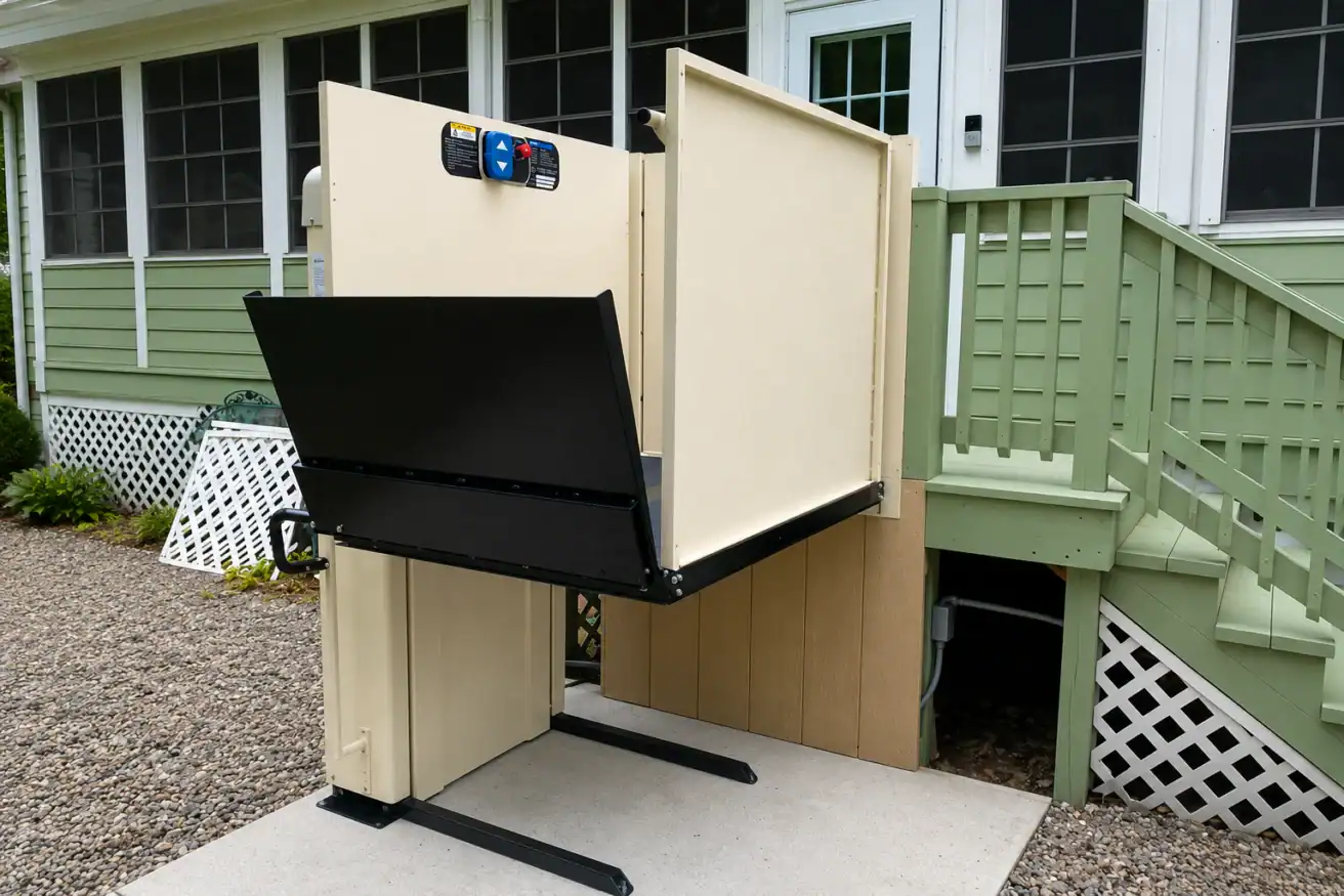A vertical platform lift is installed next to an outdoor staircase, providing wheelchair access to the porch of a green house.