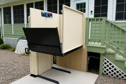 A vertical platform lift is installed next to an outdoor staircase, providing wheelchair access to the porch of a green house.