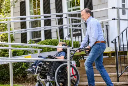 A man pushes a child in a wheelchair up a metal accessibility ramp outside a house with beige siding and black shutters.