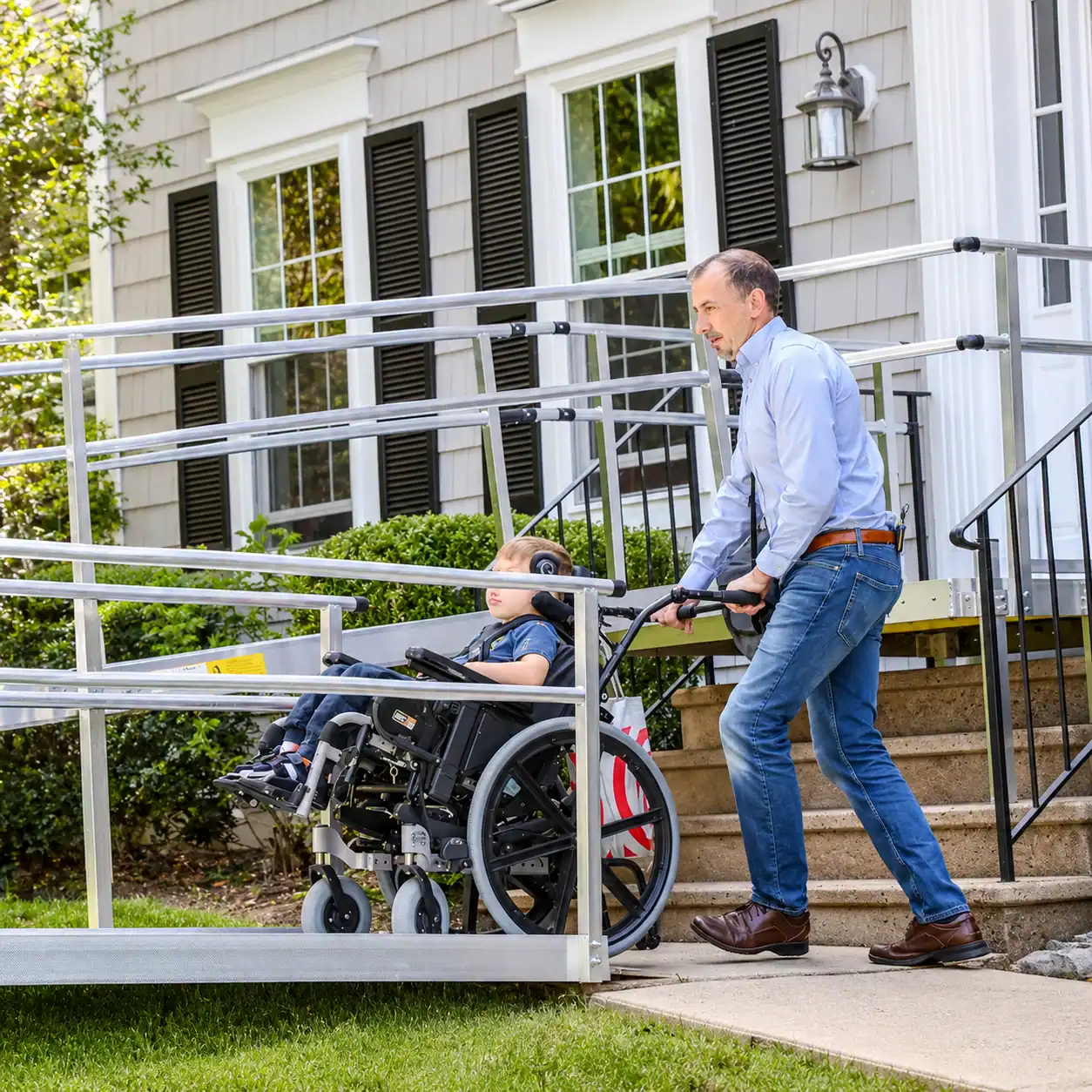 A man pushes a child in a wheelchair up a metal accessibility ramp outside a house with beige siding and black shutters.