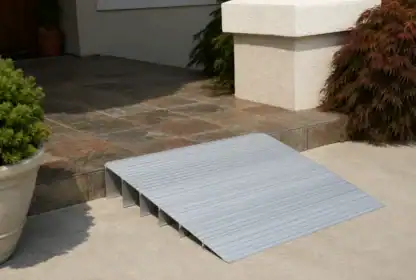 Portable metal wheelchair ramp placed on a step, connecting a tiled entryway to a concrete walkway, with plants nearby.