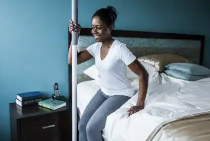 Woman sitting on the edge of a bed, holding a support pole with one hand, with books and a phone on the bedside table.