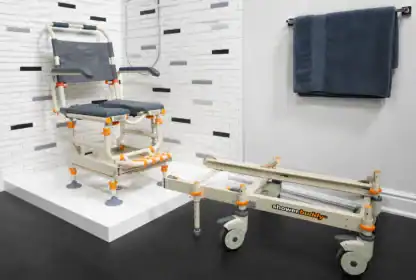 A shower wheelchair with orange accents stands on a platform in a tiled bathroom, with a matching transfer base and a navy towel hanging on the wall.