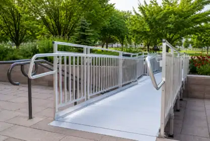 A metal wheelchair-accessible ramp with handrails on both sides, set in an outdoor area with paved ground and green trees in the background.
