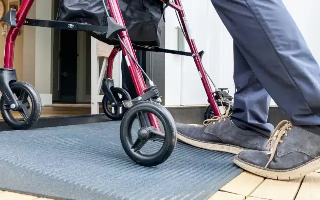 Person with navy pants and tan shoes standing next to a red walker on a textured mat by a doorway.