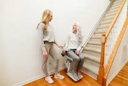 Two women are by a staircase; one is seated on a stair lift, and the other stands beside her, both dressed casually in light-colored clothing.