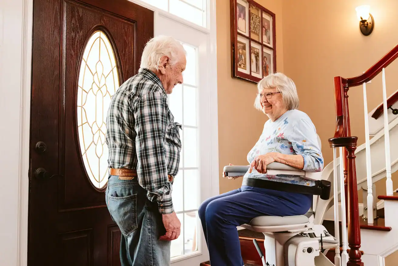 An elderly woman sits on a stair lift while an elderly man stands nearby in a well-lit home entryway next to a staircase.