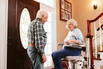 An elderly woman sits on a stair lift while an elderly man stands nearby in a well-lit home entryway next to a staircase.