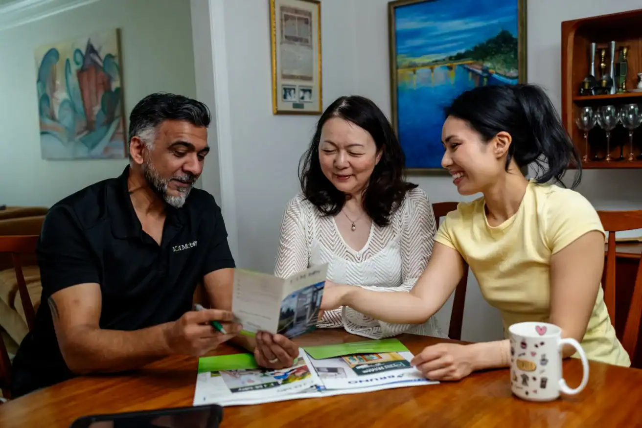 Three adults sit at a table, reviewing pamphlets and paperwork together. One person holds a pen, and another gestures toward a page. A coffee mug and tablet are on the table.