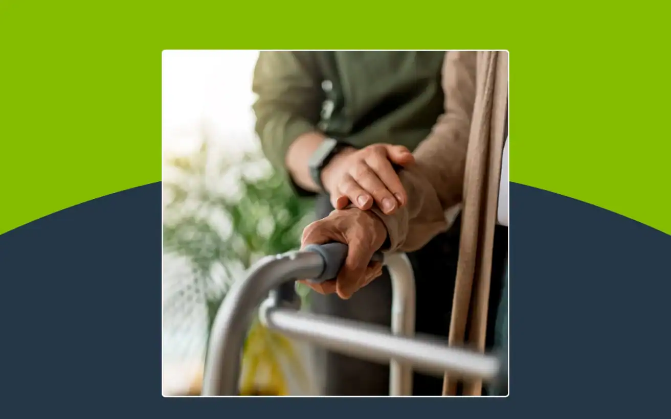 Two people’s hands rest on a walker, with one person offering support to the other. The background is blurred with greenery visible.