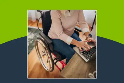 A person in a wheelchair types on a laptop at a desk in a home setting. Only the upper body and hands are visible.
