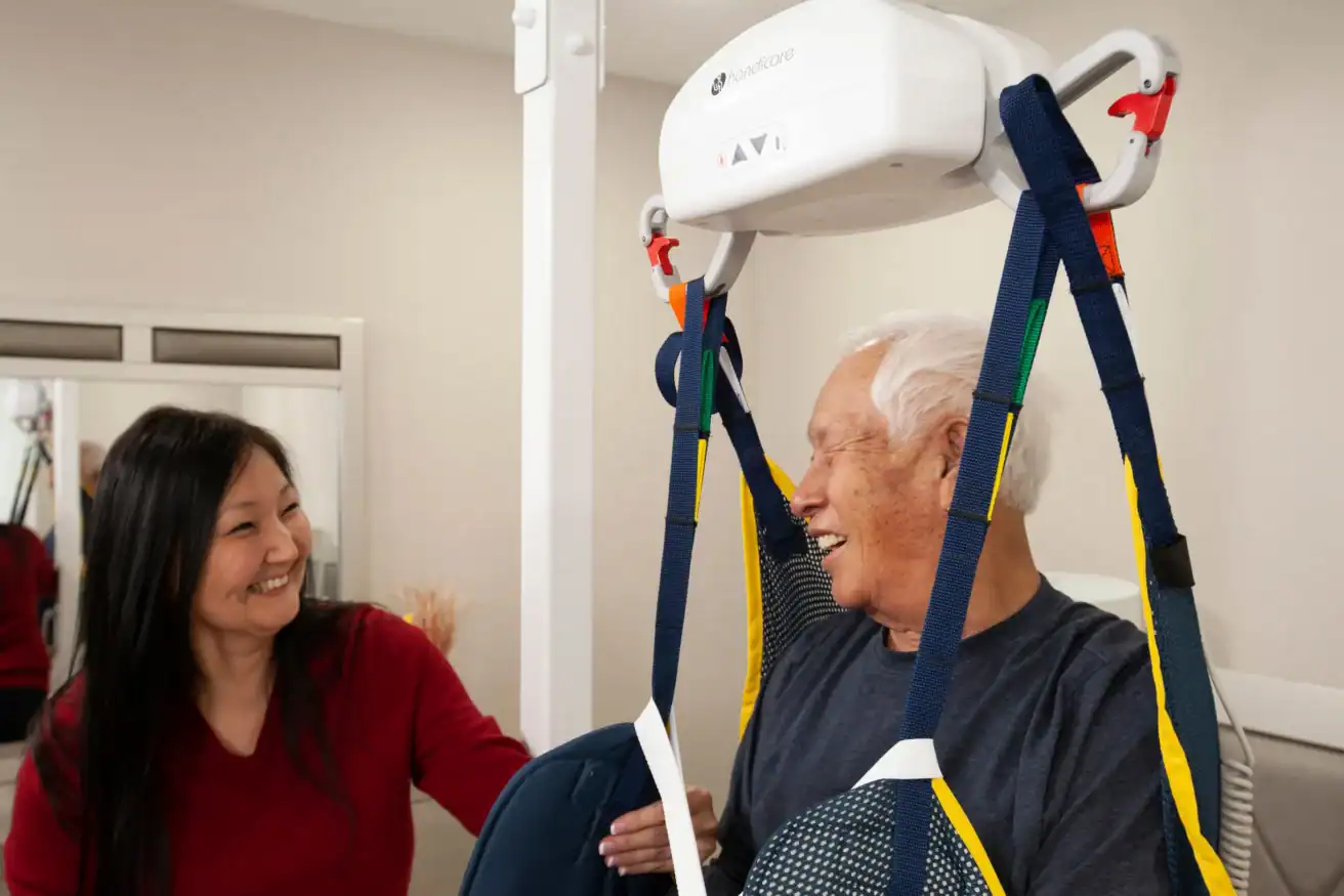 An older man in a harness lift smiles at a woman assisting him in a bright room with a mirror and medical equipment.
