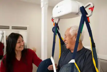 An older man in a harness lift smiles at a woman assisting him in a bright room with a mirror and medical equipment.