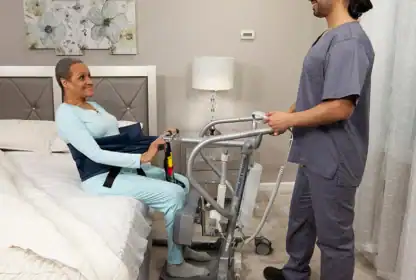 A caregiver in scrubs assists a woman using a sit-to-stand lift next to a bed in a bedroom setting.