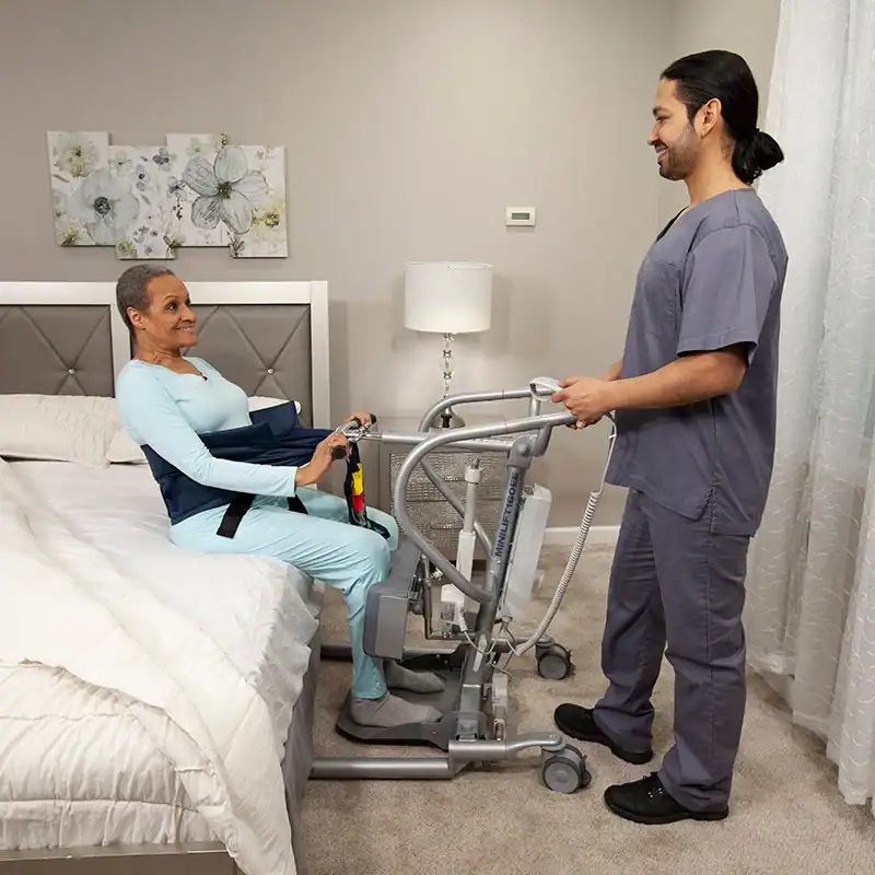 A caregiver in scrubs assists a woman using a sit-to-stand lift next to a bed in a bedroom setting.