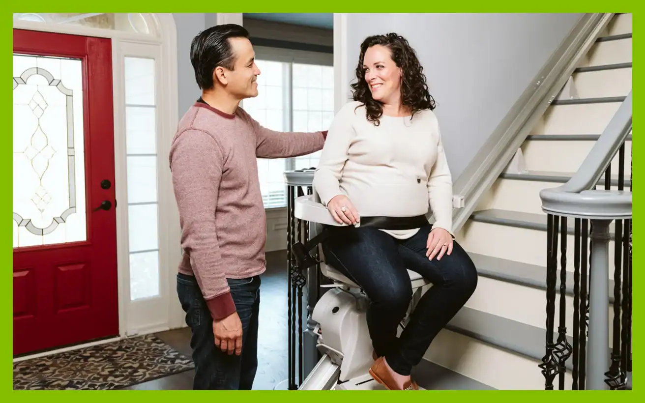 A woman sits on a stair lift at the bottom of a staircase, smiling at a man standing beside her in a home entryway.