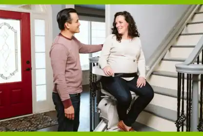 A woman sits on a stair lift at the bottom of a staircase, smiling at a man standing beside her in a home entryway.