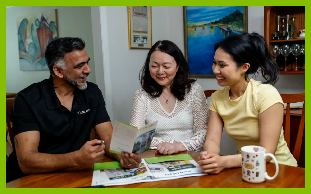 Three adults sit at a table, smiling and looking at documents together. Newspapers, a pen, and a coffee mug are on the table. Art and decor are visible in the background.