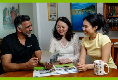 Three adults sit at a table, smiling and looking at documents together. Newspapers, a pen, and a coffee mug are on the table. Art and decor are visible in the background.