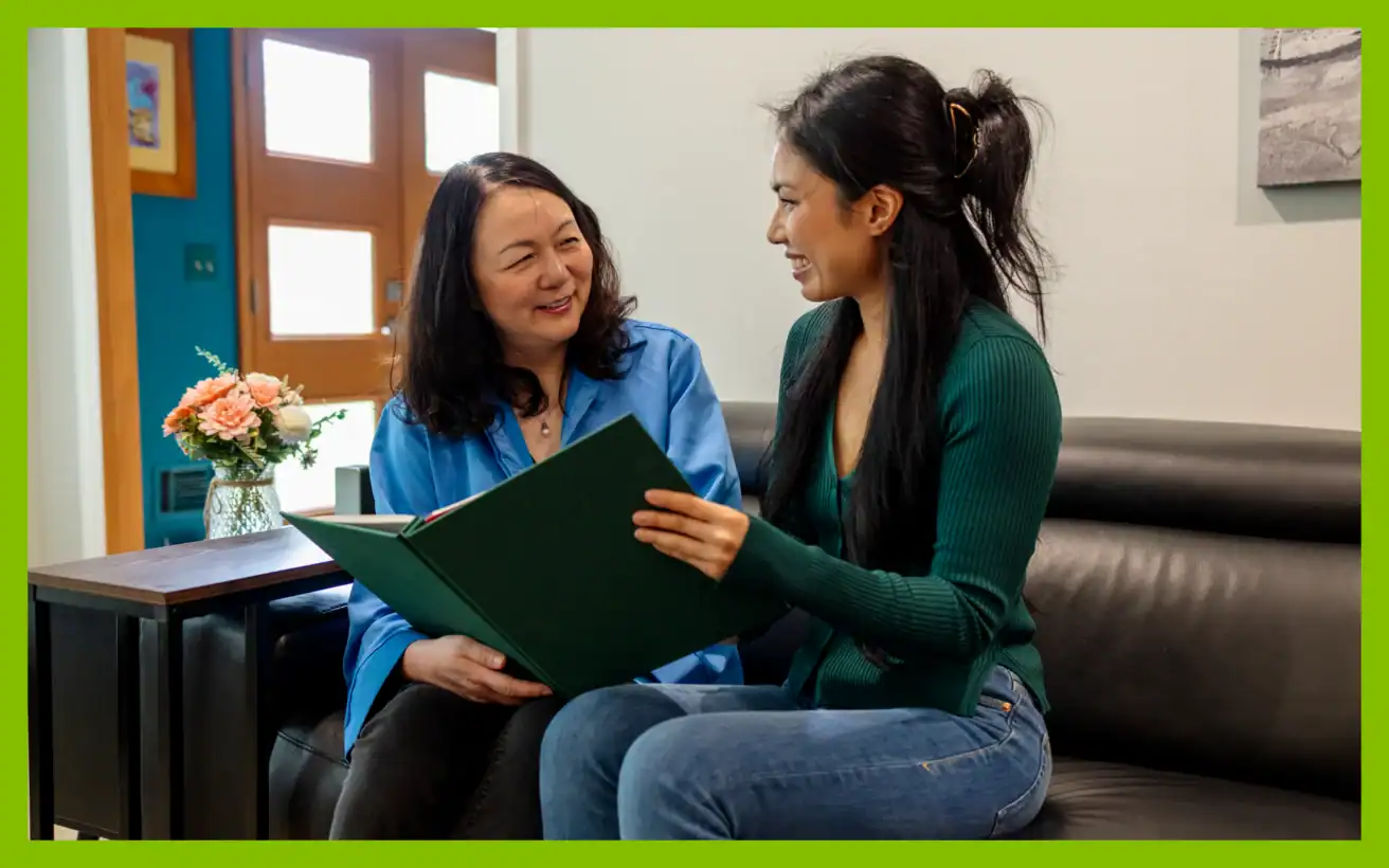 Two women sit on a black leather couch, smiling at each other while holding and looking at an open green book. A small vase of flowers sits on a table beside them.