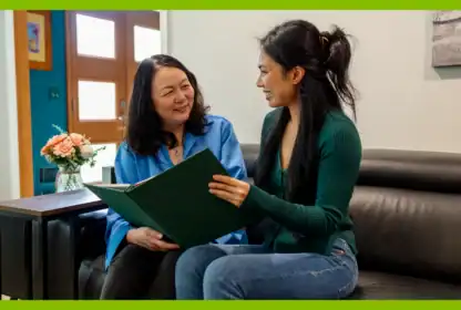Two women sit on a black leather couch, smiling at each other while holding and looking at an open green book. A small vase of flowers sits on a table beside them.