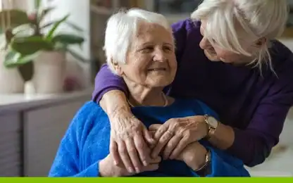An older woman in a blue blanket smiles while another woman stands behind her, embracing her shoulders and holding her hands. Both appear relaxed and content in a bright room.
