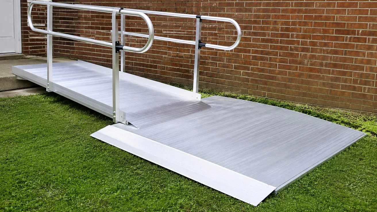 A metal wheelchair ramp with handrails leads from a concrete step to a grassy yard alongside a brick wall.