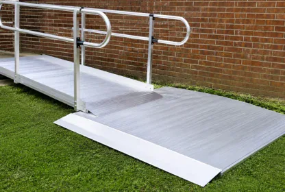 A metal wheelchair ramp with handrails leads from a concrete step to a grassy yard alongside a brick wall.