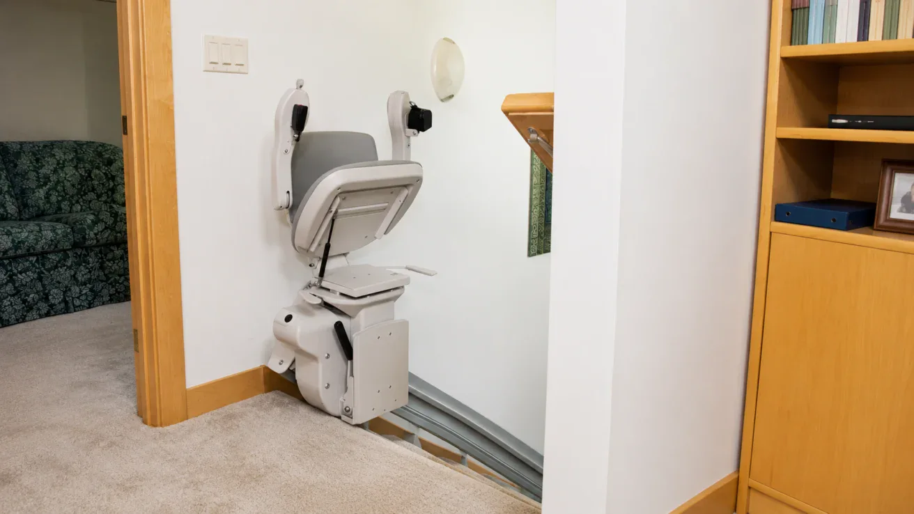 A stair lift chair is installed at the top of a staircase in a carpeted hallway next to a bookshelf and a green patterned sofa.