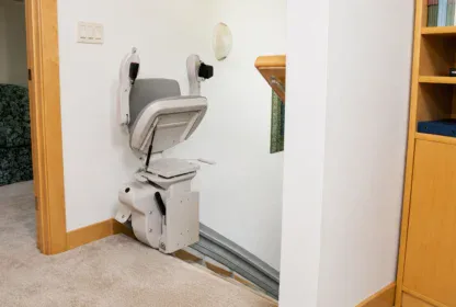 A stair lift chair is installed at the top of a staircase in a carpeted hallway next to a bookshelf and a green patterned sofa.