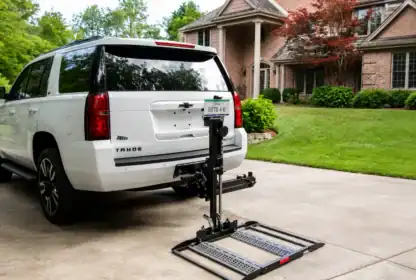 A white Chevrolet Tahoe with a hitch-mounted mobility scooter lift is parked in a driveway in front of a brick house with green landscaping.