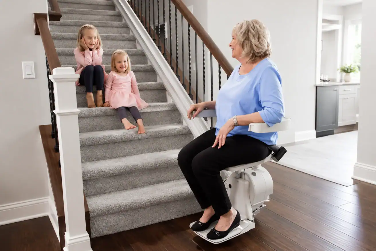 An older woman sits on a stairlift while two young girls sit on the carpeted stairs nearby in a modern home.