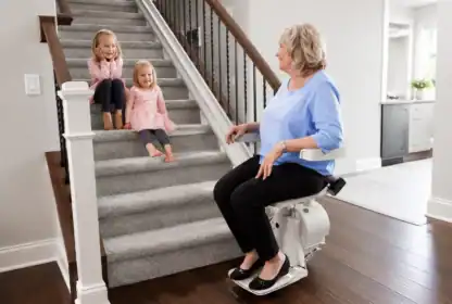 An older woman sits on a stairlift while two young girls sit on the carpeted stairs nearby in a modern home.