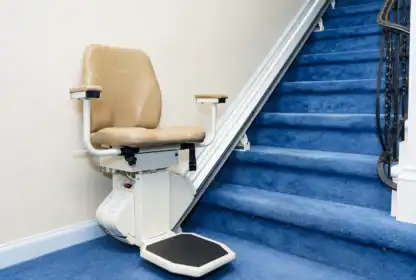 A beige stairlift is installed on a blue-carpeted staircase next to a cream-colored wall, with a black handrail visible on the right.