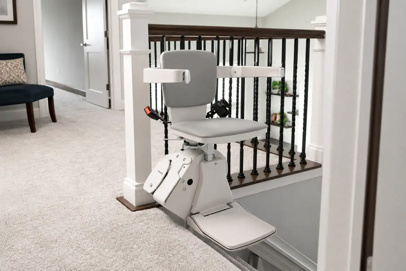 A stair lift chair is installed at the top of a carpeted staircase in a residential home, next to a banister.