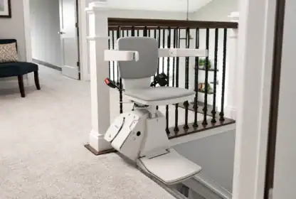 A stair lift chair is installed at the top of a carpeted staircase in a residential home, next to a banister.